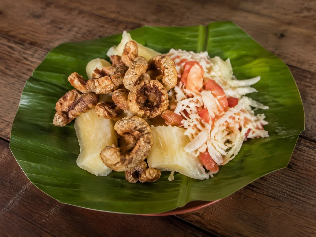 A large green banana leaf holding pale boiled cassava chunks, golden curled pork cracklings and a pile of shredded cabbage with diced tomato, on a wooden table.