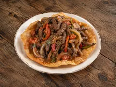 Strips of browned beef with onion, red and green pepper in glossy brown gravy piled on a large golden fried flatbread, on a rimmed cream plate on dark wood.