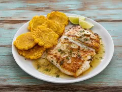 Two golden-seared white fish fillets under a glossy pale sauce with sliced garlic chips and chopped parsley, beside a stack of flattened golden plantain rounds and two lime wedges on a white plate.