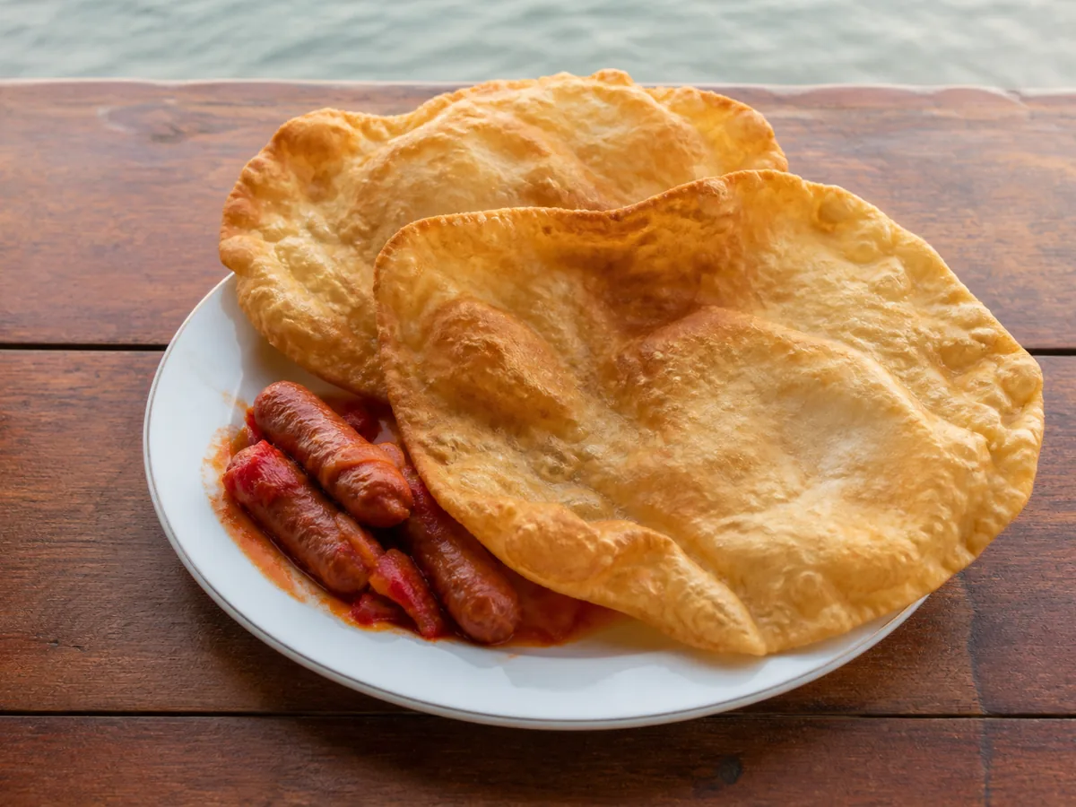 Two large golden deep-fried flatbreads with uneven blistered edges overlapping on a white plate, beside small sausages in red tomato sauce, on a wooden table by the water.