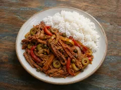 A pale ceramic plate of long shredded beef strands in orange-red sauce with sliced green olives, red pepper strips, capers and soft onion, beside a mound of white rice.