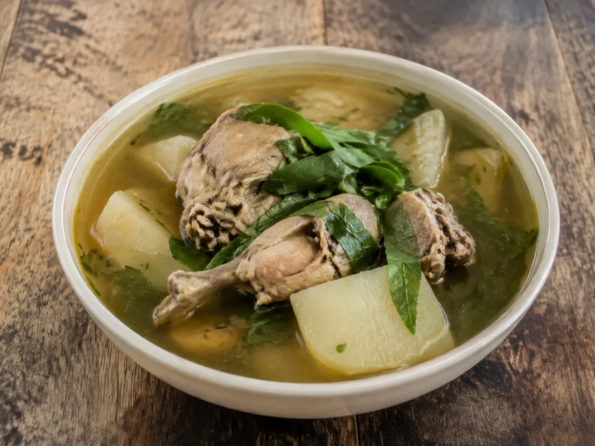 A white bowl of pale golden-green broth holding bone-in chicken pieces, chunks of white yam and long dark-green serrated leaves, on a rustic wooden table.