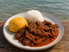 Dark beef strips in glossy crimson tomato sauce with soft onion on a rimmed plate, beside a mound of white rice and a golden corn dumpling, with the sea behind.