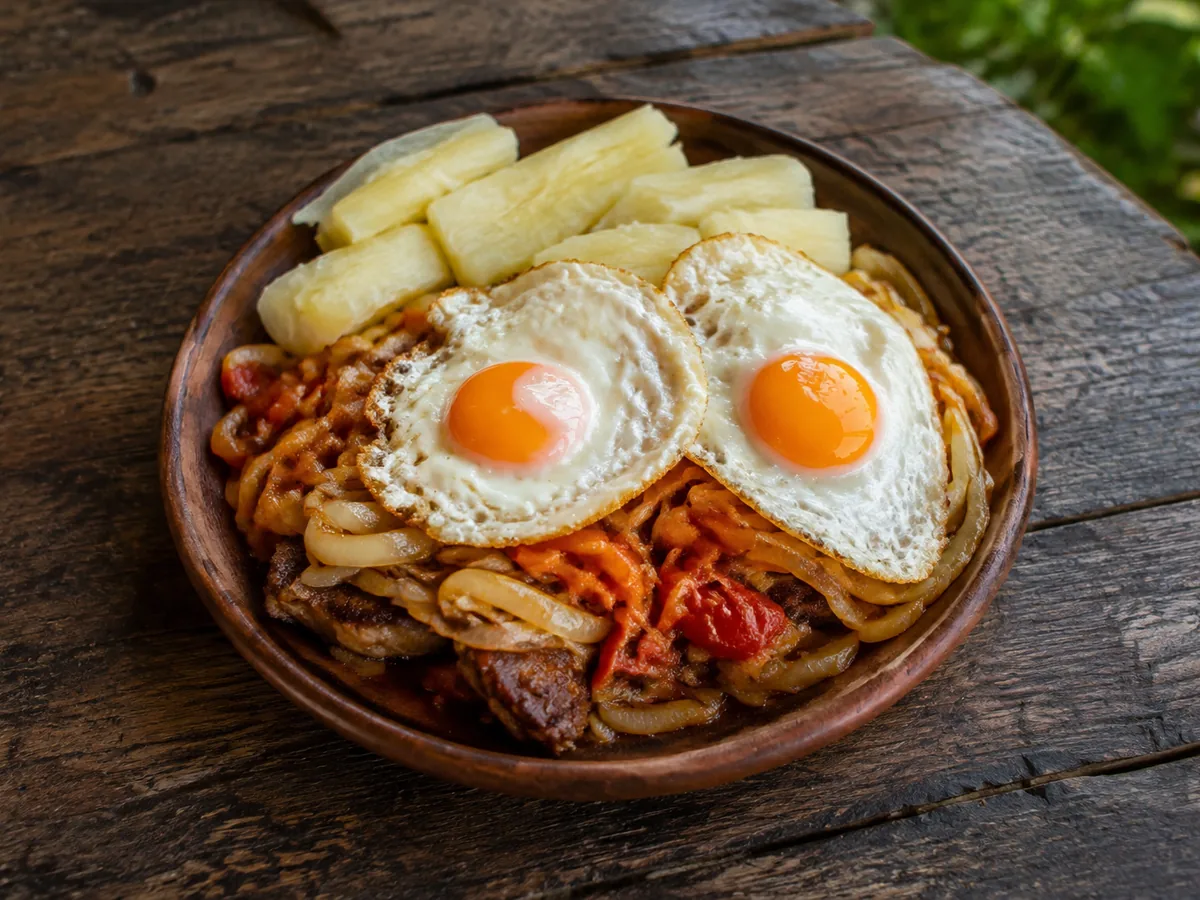 A round wooden bowl of softened onions and red tomato over browned steak, topped with two sunny-side-up fried eggs, with thick batons of pale boiled cassava tucked along the back edge, on a rough dark table.