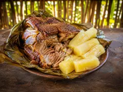 A dark clay platter lined with green banana leaves holding a slab of roast pork with dark blistered crackling and pulled shreds of meat, beside four thick batons of pale boiled cassava, with a cane fence behind.