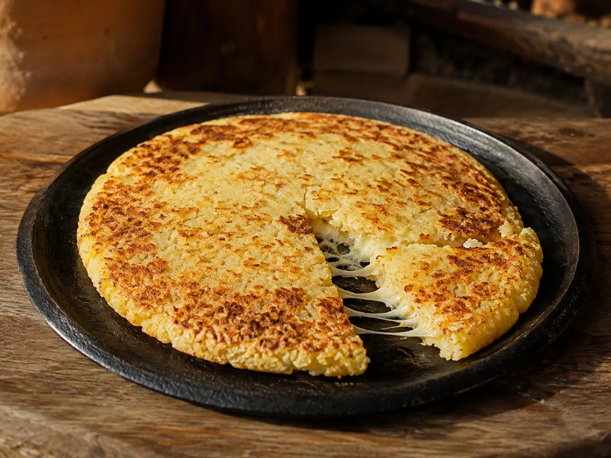 A round black iron pan holding a pale golden flatbread with a mottled toasted surface, one wedge pulled aside with fine strands of melted cheese stretching between it and the rest, on a wooden surface.