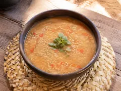 A dark clay bowl of thick pale golden-brown broth flecked throughout with fine meat particles and pieces of red pepper, topped with a small sprig of green herbs, on a woven straw mat.
