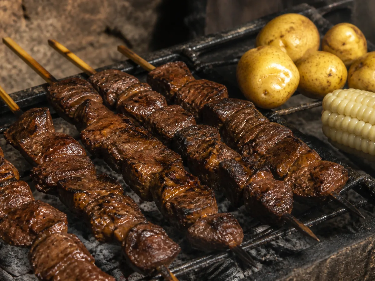 Skewers of dark, charred beef cubes laid across a sooty grill grate, with whole yellow-skinned boiled potatoes and an ear of large-kernel white corn resting on the grate behind them.