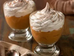 Two footed glass cups holding a layer of caramel-coloured cream under a thick swirl of pale whipped meringue dusted with cinnamon, on a dark wooden surface with a cloth and a spoon beside them.