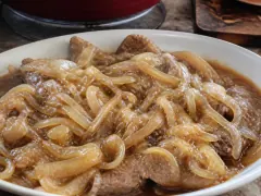 Thin steaks buried under a heap of collapsed golden onions in a caldero, sitting in dark glossy pan juices.