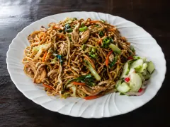 A platter of dark, glossy Surinamese bami noodles tossed with chicken, cabbage and carrot, with sweet-sour pickled cucumber on the side.