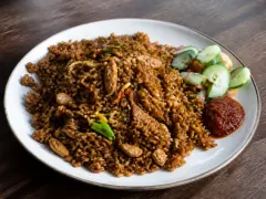 A plate of deep brown nasi goreng with pieces of roast chicken, a cucumber and tomato salad and a spoon of red sambal.
