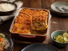 A baking dish of golden-crusted pom with one square cut away to show layers of grated pomtajer around braised chicken, with pickled cucumber and rice beside.