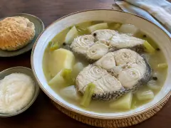 A bowl of clear broth with two skin-on grouper steaks, potato chunks, celery, onion and allspice berries, with a johnny cake and a dish of grits beside it.