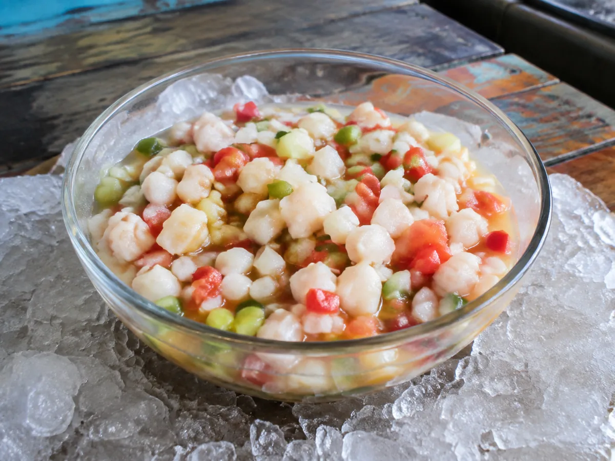A glass bowl set in crushed ice holding diced raw conch with tomato, cucumber, onion and green pepper in a pale orange citrus dressing.
