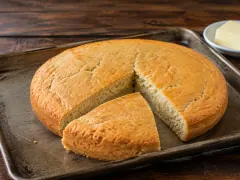 A round golden johnny cake on a baking tray with one wedge cut and pulled forward, showing a pale, dense crumb, with butter behind.
