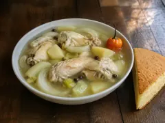 A white bowl of clear souse with chicken wings, potato chunks, onion slices, celery and allspice berries, a whole orange goat pepper on top, and a wedge of johnny cake beside.
