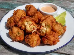A pile of craggy golden-brown conch fritters on an enamel plate, one broken open to show pieces of conch and pepper, with a dish of pink sauce and lime wedges.