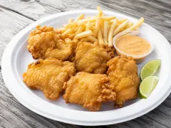 Golden fried pieces of cracked conch on a white plate with french fries, a cup of pale orange dipping sauce and lime wedges.