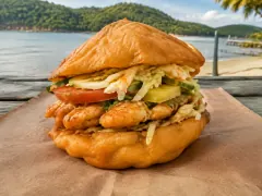A puffed golden fried bread pocket stuffed with a crisp fish fillet, cucumber, tomato and shredded slaw, on brown paper with a beach and headland behind.
