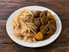 A white plate holding a mound of shredded pale flaky roti beside chunks of dark curried goat and potato in a thick brown gravy.