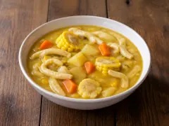 A white bowl of thick golden-yellow soup with wheels of corn on the cob, orange carrot, potato and long pale tapered dumplings floating in it.