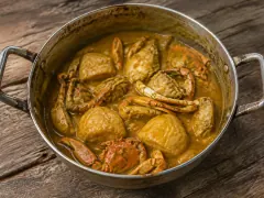 A wide metal pot of golden-yellow curry gravy holding orange-red cracked crab bodies and claws with pale flat boiled dumplings between them.