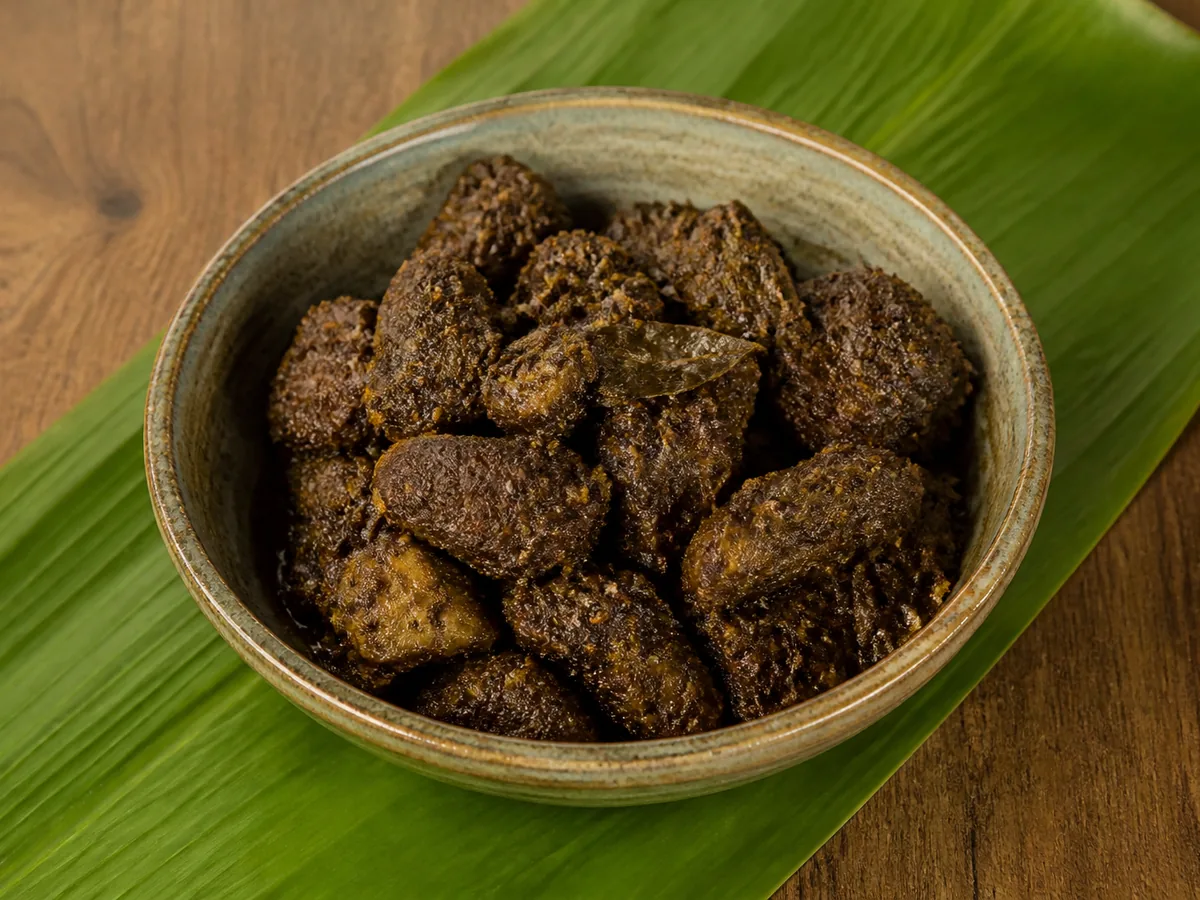 A rustic glazed bowl piled with dark mahogany-brown cubes of pork coated in a thick textured spice crust, set on a bright green banana leaf.