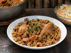 A white bowl of glossy golden-brown rice with pigeon peas, orange pumpkin cubes and two browned chicken drumsticks, topped with a thyme sprig, beside a bowl of cabbage slaw.
