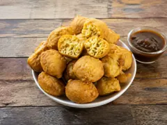 A white bowl heaped with round golden fried dough balls, two broken open to show a pale yellow spongy interior, beside a small glass dish of dark brown chutney.