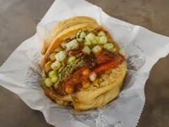 Two soft golden flatbreads holding curried chickpeas, topped with diced cucumber relish, red pepper sauce and green chutney, on greaseproof paper.
