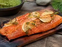 A side of ruby-red wild salmon lacquered dark amber on a smouldering cedar plank over grill grates, with thin lemon slices and dill on top.