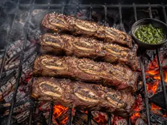 Cross-cut beef short ribs with a mahogany crust and crisp fat border on an iron grill over glowing embers, with a jar of green chimichurri beside them.
