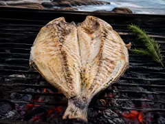 A large whole fish butterflied open on an iron grill over embers, its white flesh flecked with garlic and oregano, a rosemary sprig and lemon halves beside it.
