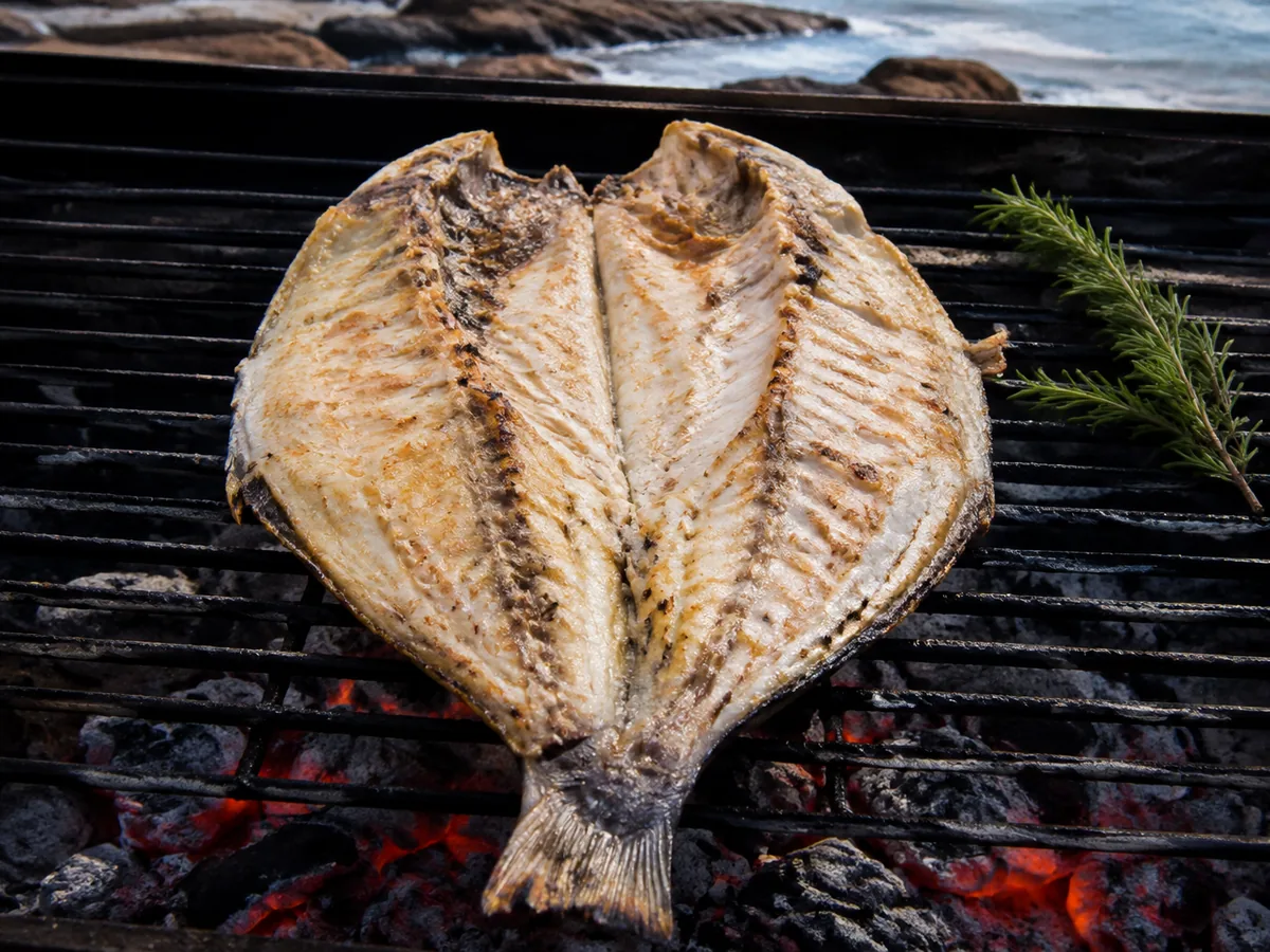 A large whole fish butterflied open on an iron grill over embers, its white flesh flecked with garlic and oregano, a rosemary sprig and lemon halves beside it.