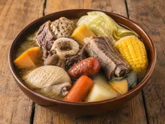 A platter of puchero: beef shank with its marrow bone, chorizo rounds, chicken, whole potatoes, sweet potatoes, pumpkin wedges and corn rounds, with a mug of clear broth beside it.