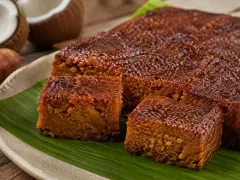 Dark mahogany squares of dense sweet potato pudding on a green leaf, with a coconut half behind.