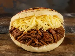 A thick pale corn cake with dark griddle scorch marks across its lid, split open and packed with long dark-brown threads of shredded beef under a heap of coarsely grated pale yellow cheese, on a rough wooden board.