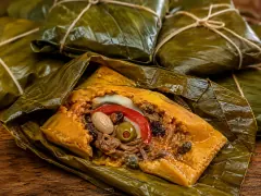 An opened parcel of deep golden-orange corn dough on a dark green banana leaf, its centre filled with shredded meat, an almond, a green olive, a red pepper strip, an onion ring, raisins and capers, with more twine-tied parcels stacked behind.