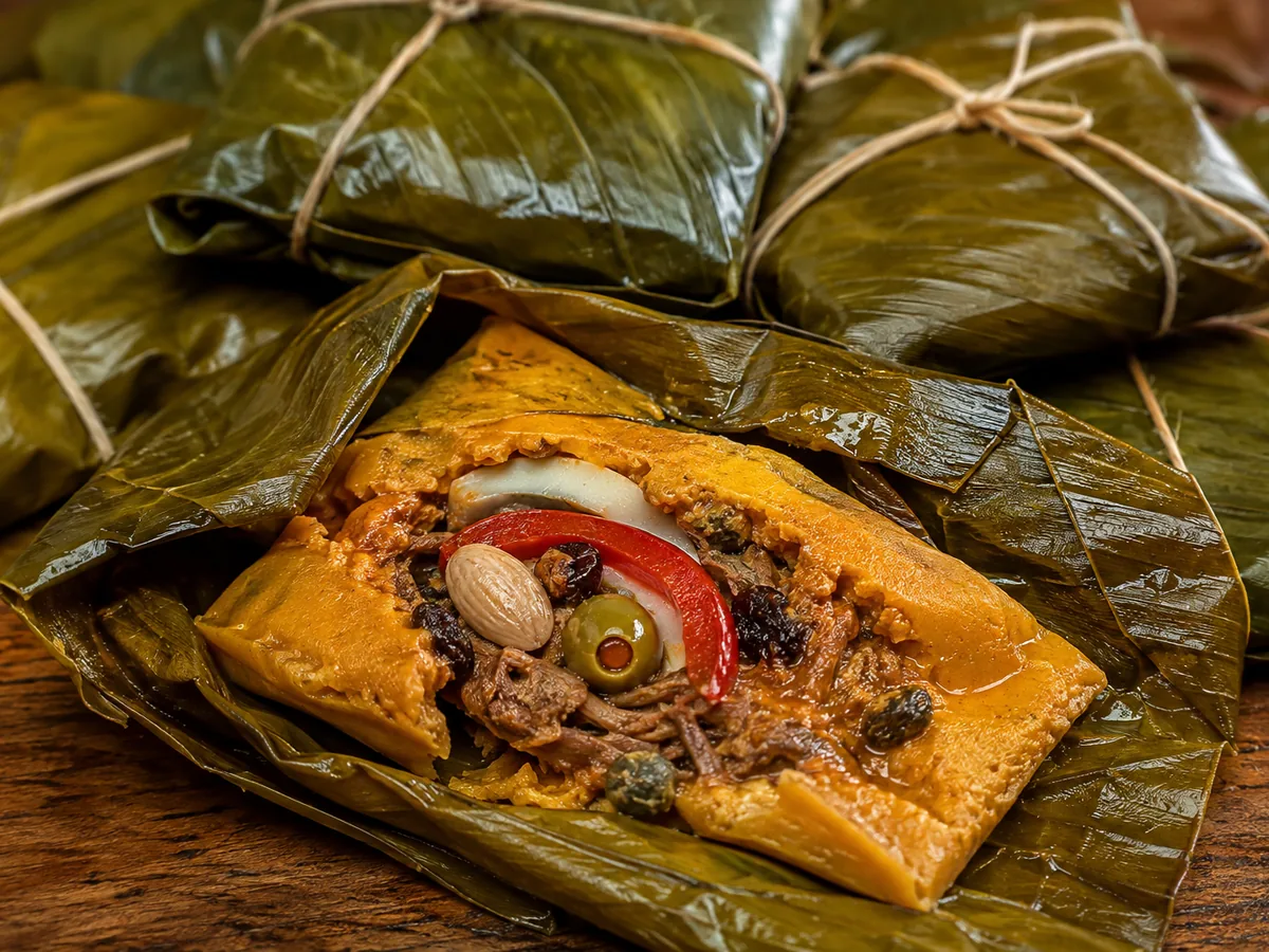 An opened parcel of deep golden-orange corn dough on a dark green banana leaf, its centre filled with shredded meat, an almond, a green olive, a red pepper strip, an onion ring, raisins and capers, with more twine-tied parcels stacked behind.