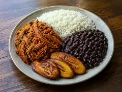 A wide stoneware plate divided between a nest of reddish shredded beef, a mound of white rice, a heap of glossy black beans and three overlapping caramelised plantain slices, on a dark wooden table.