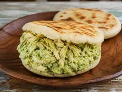 A thick pale corn arepa with dark griddle stripes across its top, split and overflowing with pale green avocado and shredded chicken filling, on a round wooden plate with a second arepa behind it.